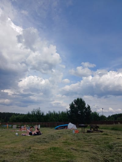 The image depicts an outdoor scene at a recreational area, likely a park or campground, near the city of Revda, Russia. A group of people are scattered across a grassy field, some reclining on blankets, others engaged in conversation. In the background, a line of trees forms a natural border, with a wooden fence running along its base.
Several objects are visible in the foreground and midground. A tent, several kayaks or canoes, a picnic table with benches, and a soccer ball are present. The sky above is filled with dramatic clouds, suggesting varied weather conditions. The overall impression is one of a relaxed, leisurely gathering in a natural setting.