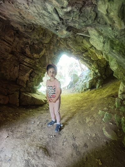 A young child, wearing a pink t-shirt with a wave graphic, pink pants, a dark baseball cap, and blue sandals with white socks, stands inside a cave. The cave walls are rough and rocky, with some areas covered in moss. The floor is dirt and pebbles. The child is looking towards the camera with a surprised expression. Sunlight streams in from the cave entrance in the background, illuminating the scene. This cave is located in Teplice nad Bečvou, Czechia, a region known for its karst landscape and numerous caves.