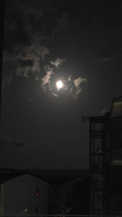 An overcast night sky is illuminated by a bright full moon, partially obscured by scattered clouds. In the foreground, the silhouettes of buildings are visible, with the right side of the frame dominated by a modern structure featuring a dark, multi-story facade with visible vertical supports and a cantilevered roof. Below this structure and to the left, the shape of a residential building is discernible, with a faint red light glowing from one of its windows. The horizon line shows the dark silhouettes of distant hills. The image quality is grainy, suggesting it was taken with a low-light setting. No people are clearly visible. The location is the city of Viterbo, Italy, at night.