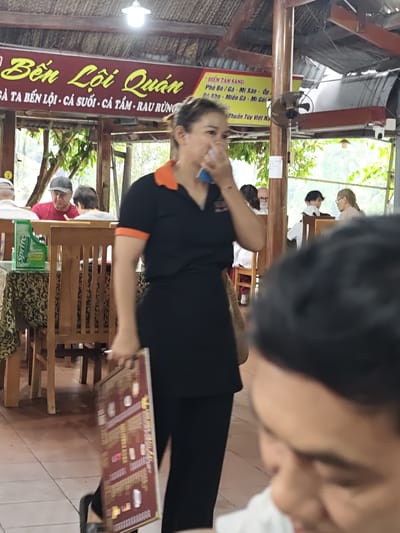 A medium shot captures a woman in a black uniform with an orange collar standing and holding a red menu board. She is positioned in the foreground, slightly off-center to the right. Behind her, in the mid-ground, several people are seated at wooden tables. A sign above the seating area reads "Bến Lội Quán" in yellow lettering on a dark red background. The establishment appears to be an outdoor or semi-outdoor restaurant with thatched roofing. In the background, lush green foliage is visible. The overall scene suggests a casual dining environment in Nam Khanh Vinh, Vietnam, possibly a rural or riverside eatery given the name "Bến Lội" which translates to "Lội River" or "River Landing."