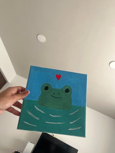 A hand holds a square canvas painting. The painting depicts a stylized green frog with a black smile and eyes, and a red heart shape. The frog is partly submerged in water, with the left side of the canvas a solid blue, representing the sky. The right side depicts darker green water with vertical white lines. The background includes a white ceiling with two circular lights, and a portion of a black television screen.