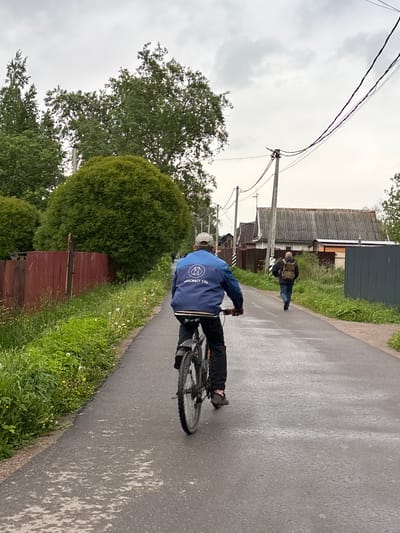 A person on a bicycle is riding on a paved road, away from the viewer. The person is wearing a blue jacket with a logo on the back and a light-colored hat. In the background, another person is walking along the side of the road. Houses and vegetation line the road. The location appears to be in the city of Ulyanovka, Russia.