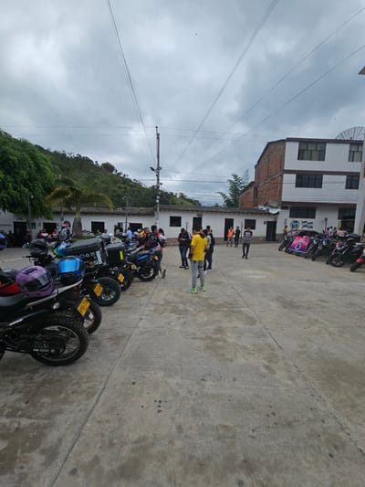 The image depicts an outdoor concrete plaza or square in Bucarasica, Colombia, under an overcast sky. A large group of people, primarily men, are gathered, with numerous motorcycles parked on both sides of the plaza.

On the left, a row of motorcycles is parked. The closest motorcycle has a purple helmet resting on its seat, and its license plate reads "VYO 77H." Another visible license plate is "XJC 00H." Many of the motorcycles are dark-colored, equipped with rear storage boxes, and feature knobby tires. A large green tree and a palm tree partially obscure a low, white building with dark doors and windows. Behind it, a longer, white building with a brown roof and arched features is visible.

On the right side of the plaza, more motorcycles are parked. A large purple and white banner, displaying "I ANIVERSARIO" (First Anniversary) and a graphic logo, is draped across two of these bikes. Further back, against a white building, a banner featuring the flag of Bucarasica (green, white, blue stripes with a shield) is visible. The building on the right is a multi-story structure with a white facade, brown trim around its dark windows, and exposed red brick on its left side. A satellite dish is mounted on its roof.

Approximately 20-30 individuals are present in the plaza. A man in a bright yellow t-shirt, light grey pants, and bright green shoes stands prominently in the foreground, facing away from the viewer. Other individuals are seen standing, conversing, or observing their surroundings. One man wears a black and white long-sleeved shirt with a graphic design, standing near the motorcycles on the left. Another person in an orange safety vest is visible towards the back right.

The background features lush green hills covered in dense foliage under the grey sky. Multiple power lines crisscross the upper portion of the frame, and a utility pole with light fixtures stands on the right. The concrete ground is a light grey, showing various stains and darker spots, with visible expansion joints.