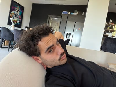 A male individual with dark, curly hair, a mustache, and stubble is depicted lying on his back on a light-colored sofa, looking directly at the camera. He wears a black long-sleeved garment, with his left arm bent and positioned behind his head or resting on the sofa. The scene is an interior living space, identified as being in the city of Mougins, France. The background features white walls contrasted with a dark grey accent wall. To the left, a dining area includes a dark table with black chairs and a vase containing a bouquet of red flowers, situated beneath a framed art piece depicting a crowned bulldog in a graffiti style. To the right, a kitchen area is visible, featuring a large dark refrigerator with a water dispenser and colorful magnets, dark overhead cabinetry, and a window or glass door providing natural light. A prominent light-colored structural pillar is located in the mid-ground, separating parts of the living space, and a multi-tiered wooden shelf unit with various objects is visible beyond it to the far right.