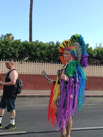 The image depicts an outdoor scene in San Bartolomé de Tirajana, Spain, on what appears to be a sunny day. A person dressed in elaborate, colorful attire associated with Pride parades or festivals is the central focus. The individual is wearing a large headdress made of rainbow-colored feathers and an intricate silver design, along with a matching flowing garment. They are holding a small rainbow fan.
To the left, a man in casual wear (tank top, shorts, sneakers) walks along the sidewalk, carrying a backpack. The background features a low fence, a brick wall, and greenery suggesting a park or residential area. The road, marked with a white line, indicates the scene is taking place along a street or thoroughfare.