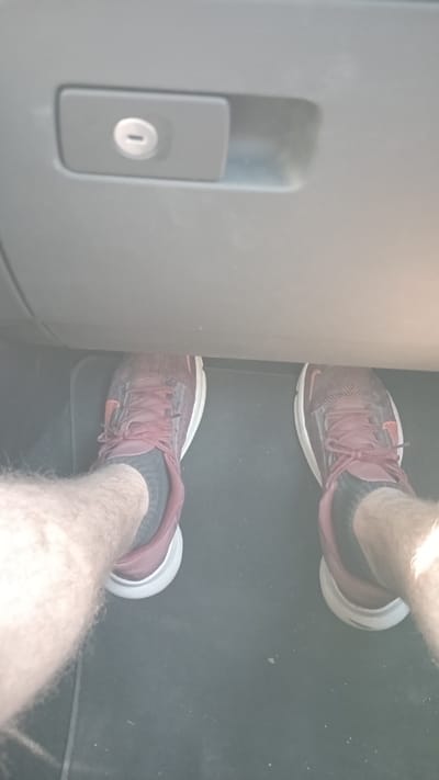 The image shows a pair of maroon and white athletic shoes, each with black socks, positioned on a dark floor. The shoes are worn by a person whose legs are visible from the knees down, covered in light-colored hair. Above the shoes, a grey dashboard is visible with a small, metallic lock mechanism. The scene appears to be inside a vehicle. There is no information or visual content within the image that places it in Kavala, Greece.