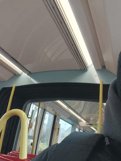 The image displays the interior of a public transport vehicle, identified as an articulated bus or tram, operating within Lingolsheim, France. The perspective is from a passenger looking forward and slightly upwards.

The ceiling features light-colored, possibly white or light grey, panels with integrated linear fluorescent lighting fixtures running along the length of the vehicle. A light blue trim panel is present beneath the ceiling structures. Bright yellow vertical grab poles are visible throughout the interior, alongside a prominent U-shaped yellow grab handle in the lower-left foreground, situated above a red surface, likely part of a seat.

A flexible, black, pleated accordion-like connector, characteristic of articulated vehicles, is visible in the middle ground, linking two sections of the transport. Large windows flank this section, offering a view of an exterior environment dominated by trees and ambient daylight. Reflections on the window surfaces show internal vehicle lights and structural components.

In the immediate foreground, the back of a person's head and right shoulder are partially visible, covered by a grey hooded garment. A black bag or backpack is situated in front of this individual. No specific actions or interactions are discernible. The scene presents a typical public transit environment in motion.