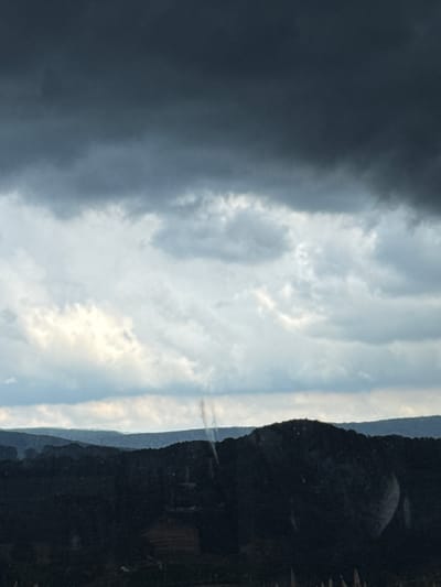 This is a vertical, slightly blurry, photographic image taken outdoors. The scene depicts a dramatic sky transitioning from dark, stormy clouds on the left to a brighter, cloud-filled sky on the right, with a dark, mountainous landscape forming the right edge of the frame.

The subjects are primarily natural elements: the sky and a mountain range. The stormy clouds are dense and dark, suggesting impending rain or a recent storm. The sky transitions to lighter hues with patches of illumination, indicating that the storm is either passing or localized. The mountain range is covered in dense vegetation, appearing as a dark, textured silhouette against the sky. A thin, vertical plume of what appears to be smoke or mist rises from the mountainside in the distance, a subtle detail against the vastness of the landscape. Some dry grass or reeds are visible along the far right edge of the frame.

The image captures a moment of dynamic weather, likely during the late afternoon or early evening when light can be dramatic. The contrast between the dark storm clouds and the lighter sky creates a sense of atmospheric tension. The location cues suggest a rural or natural setting, possibly in a mountainous region. There is no visible text in the image.