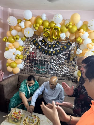 An indoor scene in a residential setting in Bidhannagar, India, depicts a 50th-anniversary celebration. The background features a large balloon arch in white, gold, and peach, centered with a silver metallic square-patterned foil backdrop. Gold foil balloons forming the number "50" are prominently displayed, with a black banner below reading "HAPPY ANNIVERSARY" in gold letters. An elderly bald man in a light blue shirt and dark pants, and an elderly woman in a green patterned saree, are seated on a brown sofa, jointly cutting a round white cake with a golden "50" topper. A floral arrangement in a clear box is visible on the table next to the cake. Several other individuals are partially visible on the right, observing the couple, with at least two individuals clapping.