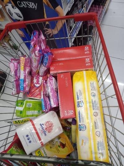 A shopping cart, observed from above, is filled with various household and personal care items in a retail establishment in Nganjuk, Indonesia. The cart, featuring a silver metal frame and red plastic handlebars, contains multiple products. Prominently visible are three red rectangular boxes, two stacked on top of each other, bearing the "SCM" logo and "6-12" designation, with two also indicating "1Kg." Adjacent is a large yellow package, likely containing diapers, with "12 JAM" text. Scattered throughout are numerous small pink plastic sachets, some labeled "So Soft" and "Rp 1.000 x 2" or "BELI 3 GRATIS." A white package of "Selection facial cotton" with a red floral design rests on top of a green package, with the brand "Milna" partially visible. Another yellow package featuring a teddy bear and fruit illustration is also present. The background shows a light-colored tiled floor, merchandise shelves, and a large promotional poster depicting a male model in a black and yellow shirt with "GATSBY" branding and a soccer ball motif.