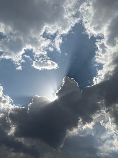 This is a low-angle, vertical shot of a dynamic sky above Kaloyanovo, Bulgaria. Dramatic, dark clouds fill most of the frame, creating a sense of foreboding. However, a radiant sun is breaking through, casting powerful rays of light that pierce the gloom. The sunbeams fan out from behind a large, shadowed cloud formation, illuminating wisps of lighter clouds and creating a striking contrast between light and dark. The overall impression is one of nature's raw power and beauty, with the sun's light fighting to emerge from the shadowed sky.