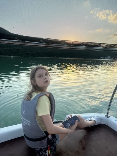 In the city of Abrau-Dyurso, Russia, a young girl with blonde hair tied in a ponytail sits on a boat, looking over her shoulder towards the camera. She is wearing a yellow t-shirt and a grey life vest with white writing and a logo on the back. The girl holds a smartphone in her left hand. Her legs are extended, and she is wearing whiteCrocs. The boat is on a body of water, possibly a lake or reservoir, with calm waves reflecting the sunset. In the background, lush green hills line the opposite shore, and a few buildings are visible. The sky is a soft blue with hints of orange and yellow from the setting sun, partially obscured by a mesh screen or canopy above the boat.