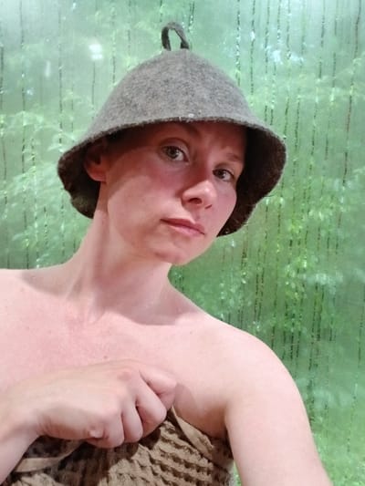 This media file shows a person wearing a grey, felted wool hat, often referred to as a banya hat. The person is wrapped in a brown towel, and their skin appears flushed and slightly damp, suggesting they may have just come from a sauna or steam room. The background is a textured, translucent green surface with horizontal lines, which could be a frosted glass door or wall, commonly found in spa or bathing facilities. The lighting is soft, and the overall atmosphere conveys a sense of relaxation and well-being. The person is looking directly at the camera with a neutral expression, their head tilted slightly. The setting appears to be indoors, likely in a private or public bathing area. There is no visible text in the image.