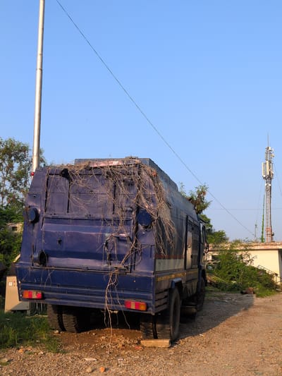 In Jammu, India, an abandoned blue armored vehicle, overgrown with dry vines, sits on a gravelly road next to a grassy area. The vehicle's rear is prominent in the frame, showing its dark blue exterior, multiple storage compartments, and red taillights. A silver flagpole stands to the left, and a cell tower is visible in the background to the right, against a clear blue sky. The scene suggests a disused or forgotten location within the city.