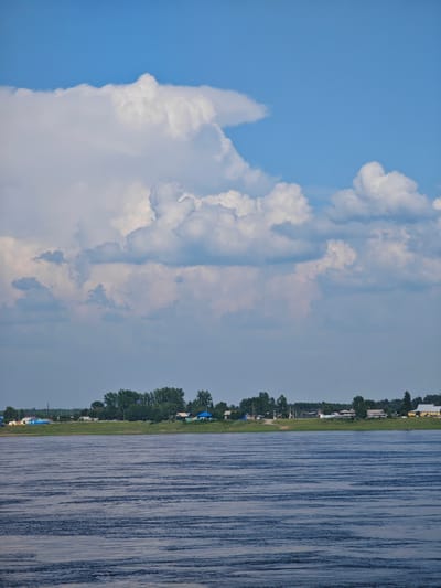 The image depicts a wide river in the foreground, with gentle ripples across its surface. In the middle ground, a green embankment is lined with trees and a village. The village consists of several small, scattered houses, some with brightly colored roofs, and appears to be situated in Verkhnepashino, Russia. The background is dominated by a vast blue sky with large, fluffy white clouds. The overall scene is serene and natural, suggesting a peaceful rural setting.