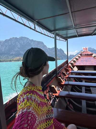 The image shows a person in a black baseball cap on a boat with turquoise water and mountains in the background. The person is wearing a multicolored mesh top in shades of yellow, orange, white, and pink. The boat has rows of seating with black life jackets stored underneath. Another boat is visible ahead, partially obscured by the boat on which the person is standing. The environment is sunny. The image was taken in Ban Chiao Lan, Thailand.