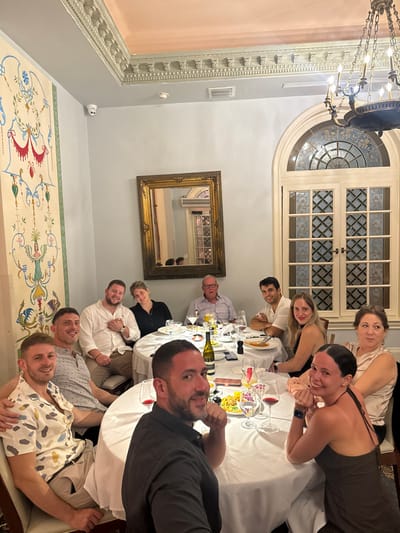 The image depicts an interior scene at Casa Tua Restaurant, located at 1700 James Avenue, Miami Beach, Florida, United States. Eleven adults, seven males and four females, are seated around a large, round dining table covered with a white linen tablecloth. Most individuals are looking directly at the camera with varying expressions, primarily smiles or neutral gazes, indicating a social gathering or dinner party.
The table is set with multiple wine glasses (red and white), water glasses, silverware, and white plates, some containing food such as pasta. At least one green bottle of white wine is present on the table, alongside condiments and other dishes.
The dining room features pale blue-gray walls. A large, ornate brass chandelier with multiple lights hangs from the ceiling, which is adorned with decorative classical molding painted in light gray and a pinkish-beige tone. On the right, a tall arched window with intricate leaded or stained glass panels and a white wooden frame is visible. On the left wall, a large, colorful mural or tapestry displays abstract floral and mythological motifs in green, red, blue, and yellow on a cream background. A large rectangular mirror with a gold ornate frame is mounted on the wall, reflecting portions of the room. A white security camera and a white smoke detector are visible on the ceiling. The lighting is warm and ambient, suggesting an evening setting.