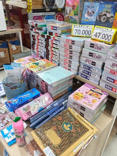 This indoor photograph captures a retail display of various stationery and school supplies, likely within a store in Sindanglaya, Indonesia. The scene is dominated by numerous neatly stacked notebooks and exercise books of different brands and designs.
In the foreground and midground, large stacks of notebooks are visible. Some have plain covers, while others feature colorful designs, including cartoon characters like a yellow duck. Price tags indicate costs in Indonesian Rupiah, such as "KIKY BUKU TULIS 70.000" and "SIDU EB 310S SPECIAL EDITION 47.000". Other visible brands and labels on the notebooks include "SIDU 58", "SIDU 38", "OKEY 40", "OKEY 89", and "FLORIST".
On the table in front, several items are arranged: a blue pencil case with a "Panda astronaut" design, a pink pencil case with a unicorn and "LOVELY HORSE" text, and a pink water bottle with a green radish-shaped top. A blue woven basket holds various pens and pencils. Another set of pens or markers are laid out next to the pencil cases. A distinctive, traditionally patterned notebook or folder with a gold and brown design is prominent in the lower right.
In the background, shelves are stocked with more products, including books with colorful covers. One book at the top right displays Korean text, "피나요 이루어". The organized presentation of items suggests a retail environment prepared for sales, possibly for the back-to-school season. No people are visible, and the indoor lighting provides no cues about the time of day or weather.