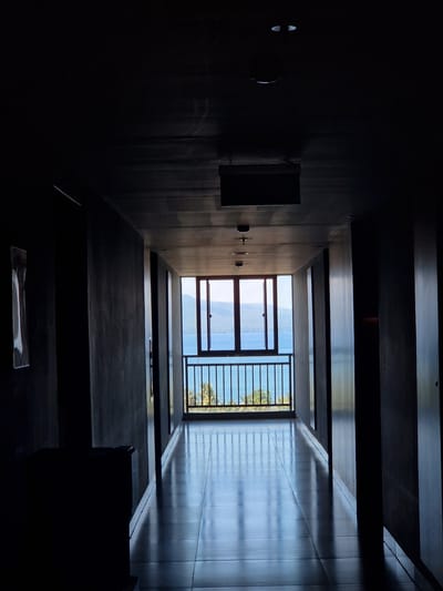 A low-angle interior shot taken in Banyuwangi, Indonesia, shows a dark, narrow hallway leading to an open area with a view of the ocean and distant land. The hallway is lined with what appears to be dark gray concrete walls and ceilings. A box-like fixture is mounted on the left wall of the hallway. At the far end, sunlight streams in through a window and railing overlooking the ocean and land.