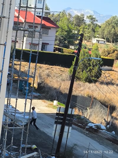 The image depicts an outdoor scene in what appears to be a residential or semi-rural area of Nicosia, Northern Cyprus / Cyprus, captured on 14/11/2025 at 12:22. The scene is illuminated by strong sunlight, creating distinct shadows.
In the immediate foreground on the left, a metal scaffolding structure, likely associated with a white-walled building partially visible, occupies the left third of the frame. Below the scaffolding, construction debris, including a yellow bucket and white sheeting, is scattered on disturbed ground.
A single adult male, wearing a light-colored long-sleeved shirt and dark trousers, is walking downhill on a narrow, unpaved path or road that traverses the midground from left to right. His body is angled slightly away from the camera, with his right foot forward. Further up the path, two green, rectangular waste dumpsters are partially visible.
To the right of the path, a dark brown utility pole with multiple attached wires stands prominently. A single street lamp fixture extends from the pole towards the left. Below the lamp, a rectangular dark sign is affixed to the pole. The path is bordered on its right by a chain-link fence, which appears partially damaged, separating the path from a steep, dry, and overgrown hillside. Piles of white bags, possibly containing waste or materials, are visible near the base of the fence on the slope.
In the background, a multi-story residential building with a red tiled roof, light-colored walls, and numerous windows and balconies is visible behind a neatly trimmed dark green hedge. Beyond this, diverse green trees and shrubs cover undulating terrain. A smaller, single-story white building with a darker roof is situated on a distant rise. In the far background, under a clear blue sky, a range of mountains with lighter, possibly snow-capped or barren, peaks is visible. The vegetation and architecture are consistent with the Mediterranean climate of Cyprus.