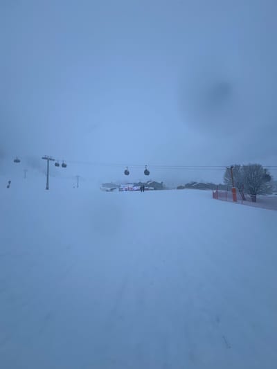 The image captures a snow-covered ski resort in Val-d'Isère, France, enveloped in extremely heavy snowfall or thick fog. The scene is dominated by muted tones of blue and white, indicating low light conditions and poor visibility.
A ski lift system is a prominent feature, with cables and several chairs suspended in the air, receding into the misty distance from the bottom-left towards the top-right. Multiple dark ski lift poles or towers are visible across the snow-covered terrain.
In the mid-ground, towards the top-right, a brightly illuminated building stands out against the white backdrop. This structure, possibly a lodge, restaurant, or ski station, has visible lights, suggesting it is either evening or night, or the intense weather conditions are obscuring daylight. A few snow-laden trees are clustered near the building, and some orange safety barriers or fences are also discernible. Several indistinct figures, likely people, can be seen near the lit building, possibly gathering or seeking shelter from the elements.
The ground is uniformly covered in a thick blanket of fresh snow, with no clear tracks or distinct features due to the flat light and pervasive haze. The heavy precipitation and low clouds merge the sky and snow, creating a diffuse, almost monochromatic landscape that conveys a sense of quietude and profound winter conditions at the resort. No visible text is present in the image.