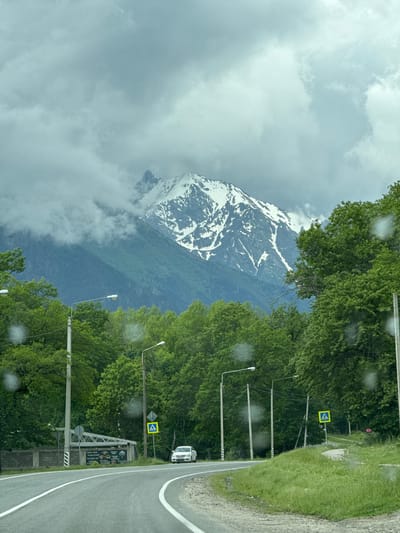 The image depicts a scenic view of a mountain range from a roadside in Verkhnyaya Teberda, Russia. A winding asphalt road with white lane markings curves through the foreground, bordered by lush green trees on either side. In the distance, a majestic snow-capped mountain rises under a cloudy sky. Several streetlights and a pedestrian crossing sign are visible along the roadside. A white car is seen driving on the road, and a small building with a patterned fence is situated on the left. The overall atmosphere is serene, with the natural beauty of the mountains and forests dominating the scene.