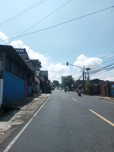 The image depicts a street scene in Zeyathiri District, Myanmar. The road is paved and has a yellow dividing line. A white line marks the edge of the road on the left. Buildings line the left side of the street, including one painted blue. Overhead power lines are visible. Several motorbikes are traveling on the road. A traffic light and a solar panel structure are positioned on the right side of the street. The sky is blue with white clouds.