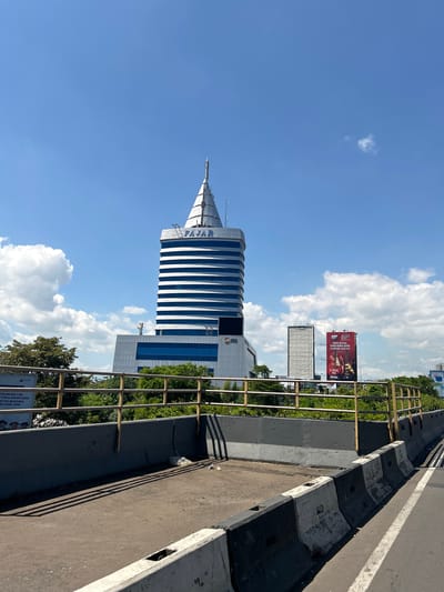This media file is a photograph taken outdoors in Makassar, Indonesia, on a bright, sunny day with scattered clouds. The perspective is from a raised vantage point, possibly an overpass or elevated road, looking down and towards a modern architectural structure. The main subject is a distinctive, conical-shaped building with a white, ribbed facade, featuring the word "FAJAR" in large blue letters near its peak. The building's design is sleek and contemporary.

In the background, other buildings and greenery are visible. A large billboard displaying a red advertisement with text in Indonesian is prominent, featuring an image of a guitar and the brand "GUDANG GARAM." The foreground shows the edge of the road or walkway, with a concrete barrier and a metal railing. The lighting suggests it is daytime, likely mid-morning or afternoon, given the clear sky and shadows. The overall scene depicts an urban environment with a mix of modern architecture and advertising. Visible text includes "FAJAR," "LPS," and details from the billboard and a sign in the lower right corner mentioning "REKTOR," "SEKRETARIS," "UNIVERSITAS BOSOWA," and dates "2026-2031."