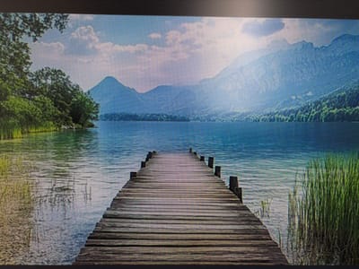 The image depicts a picturesque landscape scene featuring a wooden pier extending into a calm body of water. The pier, constructed from weathered wooden planks, leads the viewer's eye towards distant mountains under a bright sky. Lush green trees line the left bank of the water, while tall reeds grow on the right. The water reflects the sky and surrounding scenery, creating a serene and tranquil atmosphere. The scene is captured from a perspective that emphasizes the length and symmetry of the pier. There are no people or animals visible in the image. The location is not specified as being in the city of Mirnoye, Ukraine.