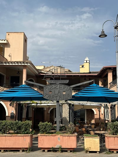 The photograph shows the outdoor seating area of a restaurant in Crabonaxa/Villasimius, Italy. Two large, bright blue umbrellas are prominently featured, providing shade over tables and chairs. A metal sign, shaped like a ship and bearing the text "LA TARTANA PIZZERIA LA TARTANA," is mounted on a pole in the foreground. Behind the umbrellas and sign, the restaurant building is visible, with a covered patio area and visible tables and chairs inside. The architecture is typical of a Mediterranean coastal town, with pastel-colored walls and tiled roofs. Planters filled with green shrubs are placed along the edge of the seating area. The sky above is partly cloudy. There are no people visible in the image.