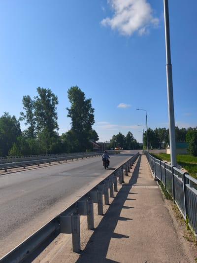 The image shows a view of a bridge with a road and a pedestrian walkway in Kady, Russia. The sky is blue with a few clouds, and the sun is shining, casting long shadows. A lone motorcyclist is traveling away from the camera on the road. The bridge has metal guardrails on both sides. Trees and buildings are visible in the background, along with a tall light pole on the right side of the image. The pedestrian walkway has a concrete surface and is separated from the road by a metal guardrail. The overall scene depicts a sunny day on a bridge in a semi-rural setting.
