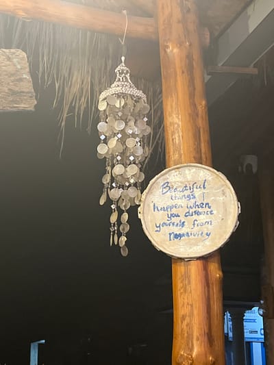 The image shows a circular object with blue writing on it, hanging from a wooden beam. The object appears to be made of a weathered material, possibly metal or plastic. The writing is in English and says: "Beautiful things happen when you distance yourself from negativity". Below this object, a decorative wind chime made of small, iridescent discs and beads hangs from a thin string. This wind chime is suspended in front of a thatched roof or ceiling. The overall scene suggests an outdoor or semi-outdoor setting, possibly a resort or a rustic dwelling, in a tropical or coastal environment given the thatched material and the context of Karimunjawa, Indonesia. The lighting suggests it could be daytime, though the background is dark, hinting at dusk or interior lighting. The focus is on the decorative items and the inscribed message, creating a calm and inspirational atmosphere.