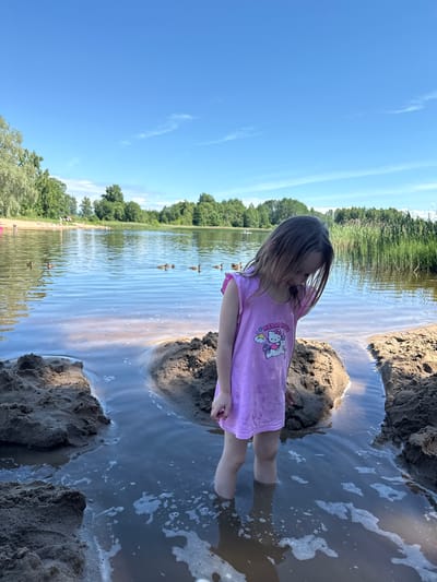 A young girl, wearing a pink t-shirt with a Hello Kitty graphic, stands in shallow water at the edge of a lake in Vaskjala, Estonia. She is looking down, and her legs are submerged up to her knees. In the foreground, there are piles of wet sand. In the background, the lake is visible, with several ducks swimming on its surface. Beyond the lake, there is a line of trees and reeds, under a bright blue sky with a few wispy clouds. Other people can be seen in the distance on the sandy shore of the lake.