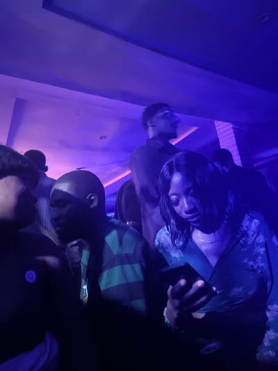 In Agwala, Nigeria, a dimly lit indoor gathering is underway, illuminated by vibrant purple lighting. In the foreground, a woman in a dark, floral-patterned top is engrossed in her smartphone, her gaze fixed on the screen. To her left, a man with a shaved head and a dark, striped garment is also present, facing towards her. Behind them, other individuals are visible, some standing and others in motion, creating a dynamic atmosphere. The environment suggests a social event or party, possibly a nightclub or a similar venue, given the lighting and the presence of multiple people.