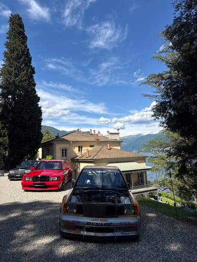 The image shows a row of vintage Mercedes-Benz cars parked on a gravel driveway at Villa Erba, Cernobbio, Lake Como, Italy. The cars, including a red AMG Mercedes-Benz, and a silver racing version with "Shell," "Dunlop," and "Roland Asch" livery, are parked in front of the villa. The racing car has its hood open. In the background, Lake Como and the surrounding mountains are visible. The sky is blue with scattered clouds, and there are trees in the foreground, framing the view. People are seen in the background near the villa.