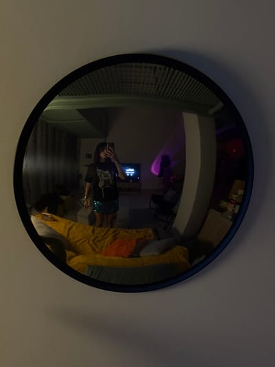 A round black-framed mirror reflects a distorted view of a room. Blinds are visible in the top-left of the mirror. A person in a black shirt and sparkly shorts holds a phone and is reflected in the center of the mirror. A TV screen and a purple light are in the background. A yellow blanket is on a bed to the right. The mirror is on a light gray wall.
