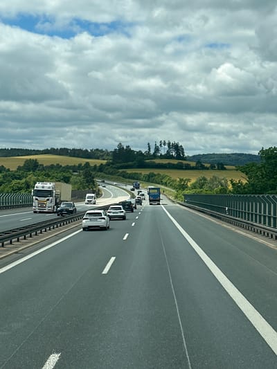 A daytime, eye-level, vertical shot shows a multi-lane highway on a cloudy day. The highway is asphalt, with white lane markers. Guardrails are visible on both sides of the road. Numerous vehicles, including cars, trucks, and vans, are traveling on the highway. The surrounding environment consists of green hills and trees, suggesting a rural or semi-rural location. The sky is filled with a mix of white and gray clouds, with patches of blue sky visible. The specific location is on the A72 highway near Weischlitz, Germany.