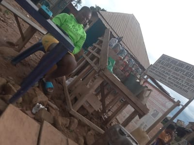 A woman in a green Balenciaga hoodie and yellow shorts sits on a blue plastic chair in Eka, Nigeria. The location is a roadside building materials store. The store's sign reads "EGEDE BUILDING MATERIAL DEALER'S & SUPPLIER". Stacks of bottled drinks are on display. The setting appears to be an open-air marketplace or trading area. The ground is made of reddish earth, with rocks and litter scattered around. The image angle is upward, indicating a low perspective.