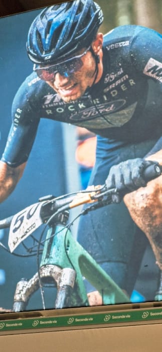 An adult male cyclist is depicted in action during a competitive event located in Vendin-le-Vieil, France. The subject, exhibiting visible exertion with a strained facial expression and open mouth, wears a dark blue long-sleeved cycling jersey. Logos "ROCKRIDER" and "Ford" are prominent on the chest, with "SRAM," "9s Seal," and "vesta" also visible. A dark blue cycling helmet with light blue accents covers his head, and dark-rimmed glasses with reddish-purple lenses are positioned over his eyes. His face, helmet, glasses, and jersey are heavily splattered with mud and water. He wears dark, full-finger cycling gloves, gripping the handlebars of a bicycle. The visible front section of the bicycle, including the frame and front suspension fork, is light green/teal and heavily caked in mud and dirt. A white race number plate displaying "50" in black numerals is affixed to the handlebars. The background is blurred, composed of dark blue and green tones, indicating an outdoor, natural, possibly wooded, and muddy environment. The bottom of the image features a green stripe with recurring "Seconde vie" text and recycling logos, which is part of the image's display medium.