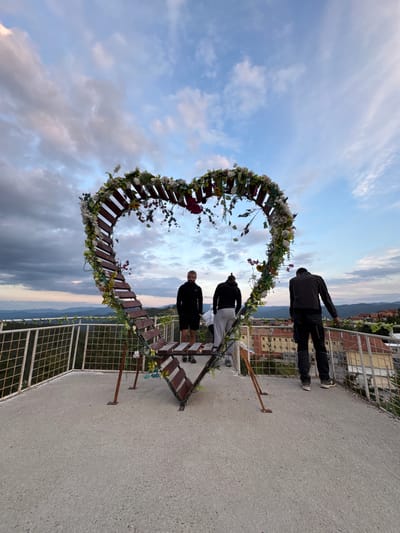 The image depicts a large, heart-shaped wooden structure decorated with artificial flowers and greenery, positioned on a rooftop overlooking a cityscape. Three individuals are present on the rooftop. One person, wearing a black hoodie and grey pants, is standing inside the heart structure, looking out. Another person, in a black hoodie and black shorts, is standing near the heart, also looking outwards. The third person, wearing a black jacket and dark pants, is leaning against a railing on the right side of the rooftop, facing away from the camera. The background reveals a valley with rolling hills and a town with residential buildings. The sky above is partly cloudy with dramatic cloud formations, suggesting dusk or a stormy weather condition. The rooftop has a concrete floor and a metal fence with a wire mesh. The heart structure is supported by metal legs. This scene is located in the city of Sveta Petka, Bulgaria.