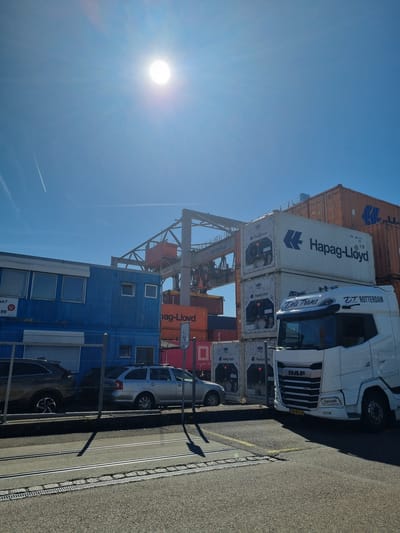 The image shows a commercial area in Saint-Louis, France, likely a port or logistics hub, based on the presence of stacked shipping containers labeled "Hapag-Lloyd" and "OCEAN NE" and the equipment needed to move the containers. A white DAF truck with "Deli Trans" and "2.7. ROTTERDAM" written on the cabin is parked next to the containers. Swiss Terminal branding is seen on the infrastructure. There is a blue, modular building in the foreground and parked cars and fence along the side of the road. The weather is clear and sunny.
