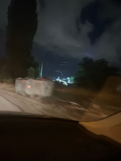A blurred, nighttime photograph taken from inside a vehicle, likely a car, shows a side view of a road and its surroundings. The vehicle's interior is visible in the foreground, with a dark dashboard and a lighter-colored passenger-side dashboard or seat creating a diagonal line across the bottom of the frame. The road itself appears wet or reflective, with indistinct streaks of light suggesting motion blur. To the left of the road, a large, dark, and tall tree is prominent. In the middle ground, a blurred, light-colored object, possibly a trailer or a large container, is situated on the side of the road, with faint red lights visible on its side. Beyond this object, there are hints of other structures and trees, including what appears to be a tall, thin pole or tower with lights. The sky above is dark and cloudy, with some lighter patches suggesting moonlight or distant city lights. The overall impression is one of a dimly lit, possibly rural or semi-urban environment captured during a journey at night. The photograph's quality is compromised by motion blur, rendering many details indistinct. The scene is in Krivina, Bulgaria.