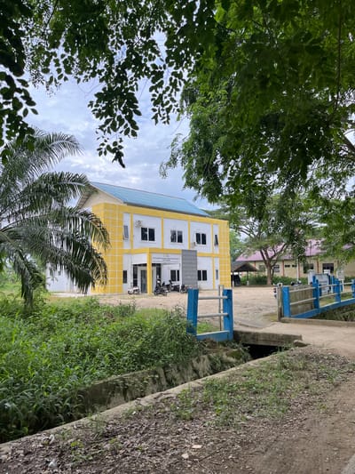 This is a rotated, outdoor, eye-level, medium shot of a building and its surroundings in Bundar, Indonesia. The building is painted yellow with white trim and has a blue tiled roof. It has multiple windows and air conditioning units. In front of the building, there are several motorcycles parked. A blue and white railing runs along the side of the building and a dirt path. To the right of the building, there is a concrete channel with overgrown green vegetation and dry leaves. The sky is cloudy, suggesting it might be an overcast day. The overall scene appears to be that of a public building or institution, possibly a school or administrative office. The time of day is indeterminate due to the lighting, but the cloudy sky suggests it is daytime. The image lacks any discernible emotional cues, but the presence of the building and motorcycles implies a functional location. Visible text on a sign above the entrance reads "PELATIHAN PEKERJA SOSIAL INDONESIA" and "BALAI PELATIHAN PEKERJA SOSIAL", indicating it is a social worker training facility.