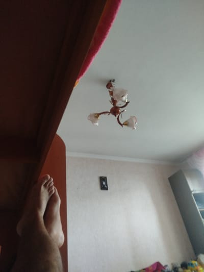 The image displays a person's bare feet crossed, resting against a dark wooden surface. Above, a chandelier with three frosted glass shades hangs from a white ceiling. The wall behind the feet is light-colored and textured, with a small dark framed picture mounted on it. A portion of a tall grey shelf unit is visible on the right side of the frame. The overall scene suggests an indoor domestic environment, likely a bedroom, with the perspective implying the photographer is lying down. The location in Bender, Moldova, is not discernible from the visual information provided.