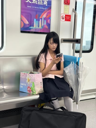 A young East Asian woman with long black hair, wearing a pink collared shirt and a dark blue pleated skirt, sits on a silver bench inside a modern subway car. She is looking down at her smartphone, which she holds in her right hand. In front of her, on the bench, is a pink gift bag with colorful flowers and text that reads "BEST WISH THANKS YOU LOVE". To her right, a transparent umbrella is propped up. A large black bag rests on the floor in the foreground. The subway car features advertisements on the wall, including a purple advertisement with Chinese text and graphics of buildings and a train. Another sign above the woman's head indicates an emergency use only button. The environment appears to be clean and well-lit. The location is identified as Shuangfu, China.