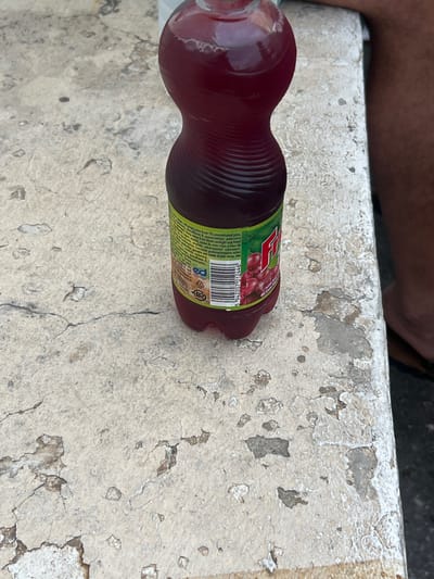 A plastic bottle containing a deep maroon liquid, identified as "Fru" brand red grape juice (barcode: 5 942207 012446), rests on a heavily weathered, light-colored concrete or stone surface in Ontinyent, Spain. The bottle's label, featuring red grapes on a green background, indicates the presence of 1% concentrated red grape juice and displays recycling symbols (PET 1). The liquid level is full, with some foam visible at the surface near the bottle's neck.

The surface beneath the bottle is predominantly off-white to pale beige, exhibiting significant deterioration, including extensive spalling, numerous fine and wide cracks, and areas where the top layer has chipped away to reveal darker, possibly dirt-filled, sub-layers. The texture is rough and irregular.

To the right of the bottle, a portion of a human leg and foot is visible. The skin tone is dark, and the person is wearing a dark flip-flop or sandal. The lower edge of a dark garment, likely shorts, is visible on the upper thigh. The leg's position suggests the individual is seated or resting adjacent to the surface. The overall environment appears to be an aged, outdoor setting.