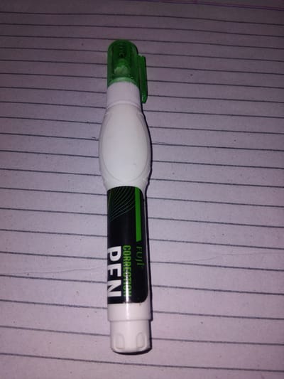 The image shows a white and green correction pen with black text on a paper with horizontal lines. The text on the pen reads "FUJI CORRECTION PEN". The pen is lying horizontally on the paper. The scene is an indoor setting, likely a desk or table, with lined paper serving as the background. There is no specific time of day or weather indicated. No people are present in the image. The object is a common stationery item used for correcting errors in writing. The location context provided is Haveli Jhangranwali, Pakistan, but this is not visually evident from the image itself. The image is a close-up shot of the correction pen, highlighting the product's details and branding.
