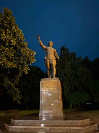 The image depicts a statue of a man in uniform, with his right arm raised. The statue is made of bronze and stands on a stone pedestal inscribed with Cyrillic text. The statue is illuminated by artificial light, suggesting it is nighttime. The background consists of trees and a deep blue sky. The statue is located in the city of Nalchik, Russia, specifically at the monument to Toraqul Bey Baibagash Baiburin. The monument is situated in a park-like setting, with a paved area and a bench visible in the lower left of the frame. The overall atmosphere is solemn and commemorative.