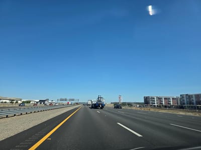 The image shows a view from a vehicle traveling on a multi-lane highway. The sky is a clear, bright blue, indicating a sunny day. The road is paved with asphalt, and there are yellow and white lane markings. On the left side of the highway, there is a gravel shoulder and a metal guardrail. Beyond the guardrail are several commercial buildings with light-colored facades and some landscaping.
In the distance, several vehicles are visible on the highway, including cars, trucks, and a blue flatbed truck carrying a large piece of equipment, possibly a generator or industrial machinery. Further down the highway, more buildings can be seen, including a multi-story apartment complex with a modern design and balconies. One building has a sign that reads "FOR LEASE." Another sign is partially visible and appears to be for "Costco."
The time of day appears to be daytime, likely late morning or early afternoon, given the brightness of the sun and the clear sky. There are no people clearly visible in the image, but the presence of vehicles suggests normal highway traffic. The scene is one of everyday travel on a suburban or urban highway.
