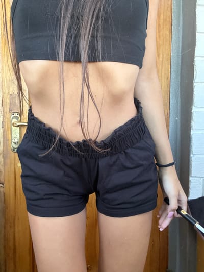 In Basarabeasca, Moldova, a person stands in front of a wooden door, their midsection and legs visible. They are wearing a black crop top and black shorts. Their long, dark hair falls forward, partially obscuring their body. The person's hands are visible at the bottom right of the frame, one holding a black and gold object, possibly a pen, and the other resting on their thigh. The background shows a wooden door with a metal handle on the left and a grey and white brick wall on the right. The lighting suggests an outdoor or well-lit indoor environment.
